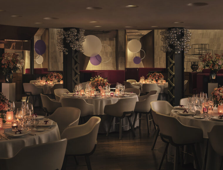 Elegant restaurant interior at night, multiple round tables set for a formal event, featuring white linens, glassware, candles, and floral centerpieces; soft lighting illuminates the room with decorative globe chandeliers and circular hanging art.